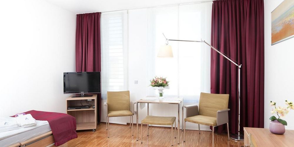 Patientenzimmer (Regelleistung) des Alexius Josef Krankenhaus. In diesem stehen zwei Stühle ein Tisch, ein Bett, ein Fernseher sowie ein Schreibtisch und eine Stehlampe. 