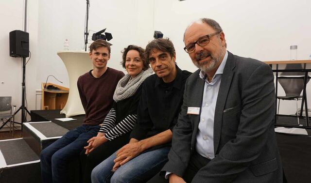 Drei Männer und eine Frau sitzen am Rand einer Theaterbühne. Alle vier schauen in die Kamera.