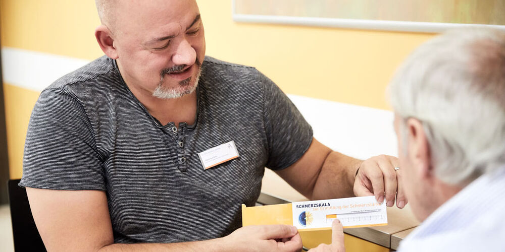 Ein Krankenpfleger sitzt einem Senioren gegenüber und hält ihm eine Schmerzkala hin. Der ältere Herr zeigt etwas auf dieser.