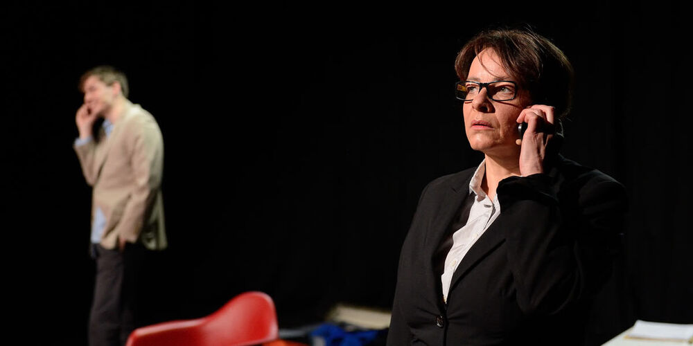 Theaterbühne auf der eine Frau im Vordergrund ist und ein Telefon ans Ohr hält. Im Hintergrund steht ein Mann und hat ebenfalls ein Telefon am Ohr. Zwischen ihnen befinden sich zwei Stühle.