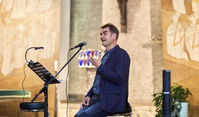 Ein Mann steht in einer Kapelle vor einem Mikrophon, spricht in dieses und gestikuliert. Schräg vor ihm steht eine Art Rednerpult.